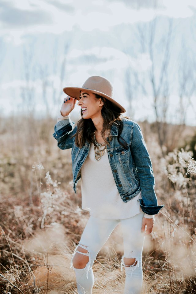 woman standing on dried grass facing right while holding her brown sun hat during daytime