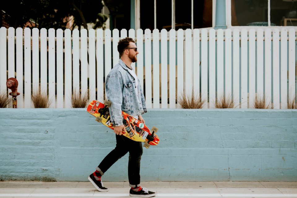 man carrying long board walking beside white fence