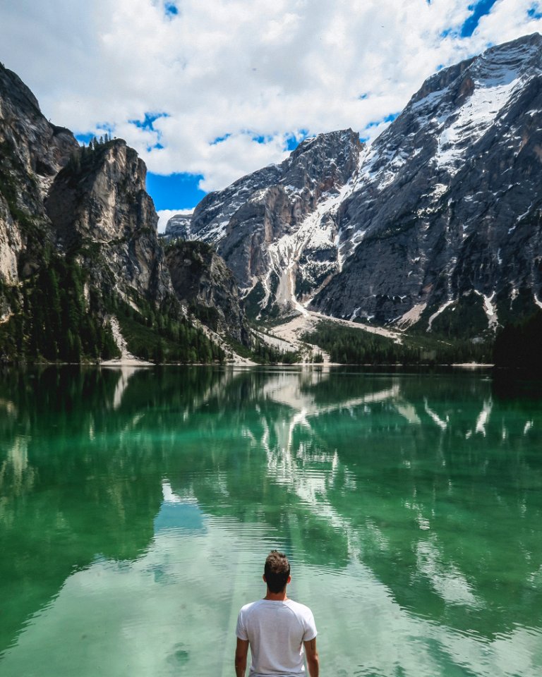 man standing in front of lake and cliff under white clouds and blue sky
