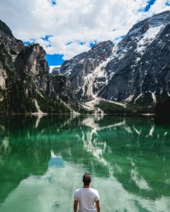 man standing in front of lake and cliff under white clouds and blue sky photo