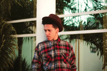 man wearing brown fedora hat and plaid dress shirt while leaning on white post photo