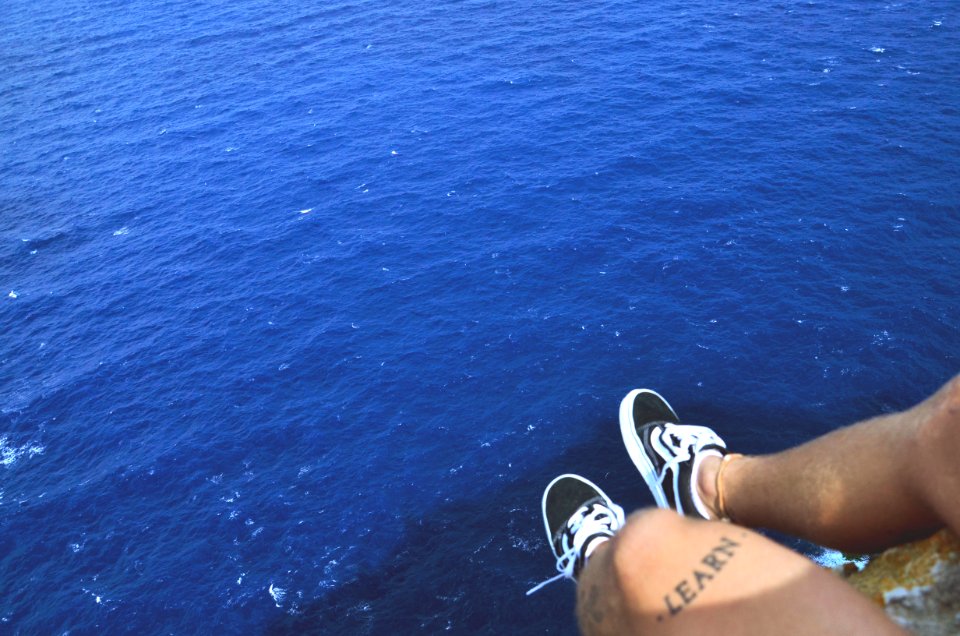 person wearing black shoes sitting on rock above water