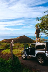man standing on roof of Jeep Wrangler parked on dirt road photo