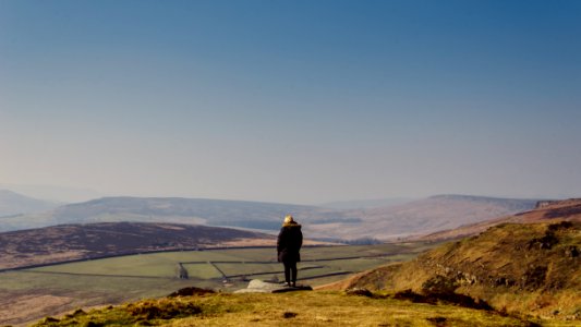 person wearing coat standing on cliff looking towards field under clear blue sky during daytime photo