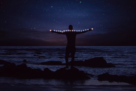 silhouette of person standing on rock on shore stretching string light at night photo
