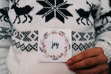 A person wearing a reindeer Christmas sweater while holding a card that says "Joy," with a wreath surrounding the writing. photo