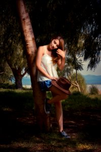 woman in white tank top and blue denim shorts leaning on brown tree during daytime