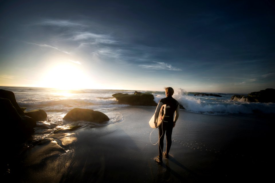 man standing carrying surfboard near seashore during daytime