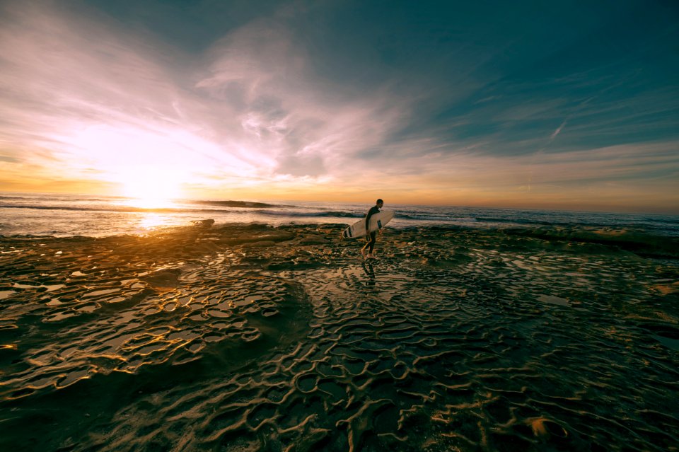 man carrying white surfboard while walking on shoreline at daytime