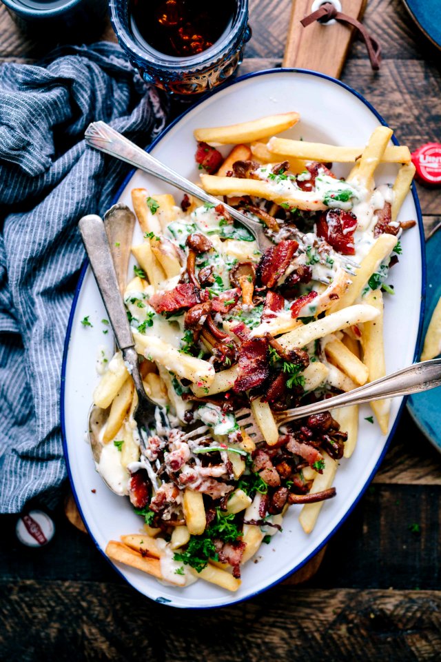 bacon strips and melted cheese topped fries on oval white and blue platter with gray stainless steel forks