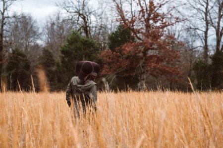 shallow focus photography of person wearing gray hooded jacket and black sun hat standing in brown grasses near green and brown leafed trees during daytime photo