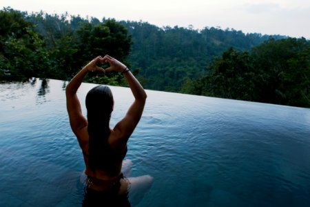 woman in infinity pool making heart hand gesture facing green leafed trees