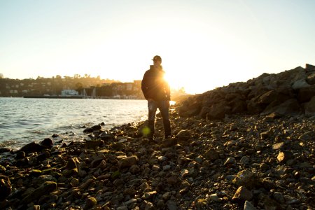 man wearing black jacket standing on stones near body of water under blue sky during golden hour photo