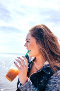 woman wearing black and grey jacket holding glass cup while drinking near body of water during daytime photo