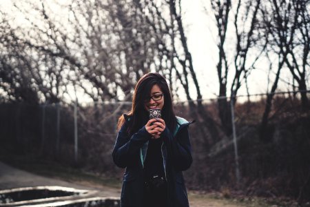 smiling woman holding smartphone standing near fence with trees during daytime photo