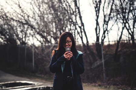 smiling woman holding smartphone standing near fence with trees during daytime