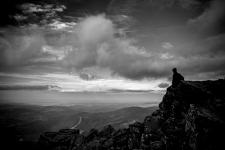 grayscale photography of man sitting on mountain peak under cumulus clouds photo