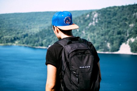 man wearing black t-shirt, black backpack, and blue fitted cap facing body of water photo photo