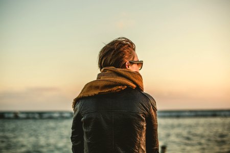 focus photo of person wearing black leather jacket with brown scarf near sea during daytime photo