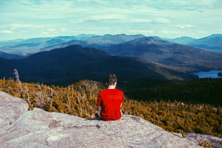 man sitting on gray rock formation looking at pine trees during daytime photo