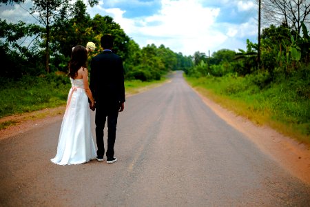 wedding couple standing on winding road between trees under cumulus cloud photo