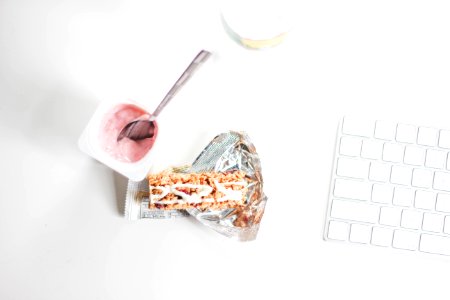 A spoon inside a small yogurt cup next to a granola bar and a Mac keyboard. photo