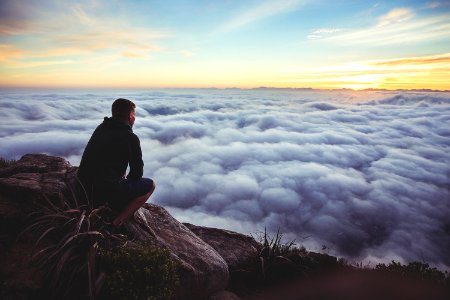 man sitting on gray rock while staring at white clouds photo