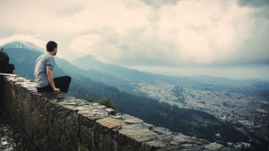 man sitting alone on concrete brick wall facing mountain and city under cloudy sky photo