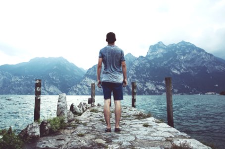 man standing on gray concrete dock facing body of water and mountains at daytime photo