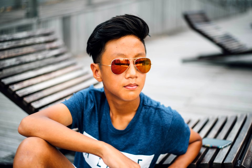 boy wearing blue and white t-shirt and sunglasses sitting near wooden lounge chair