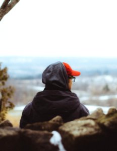 person in black hoodie and orange knit cap sitting on rock near body of water during photo