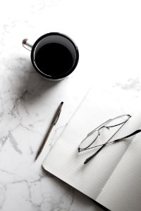 A cup of black coffee next to a notebook with a pencil and pair of glasses.