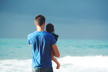 man wears blue crew-neck t-shirt holding toddler wears black hooded jacket near ocean under blue sky at daytime photo
