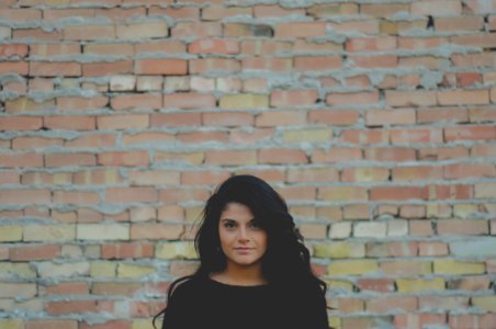 tilt shift lens photography of woman standing near brown brick wall photo