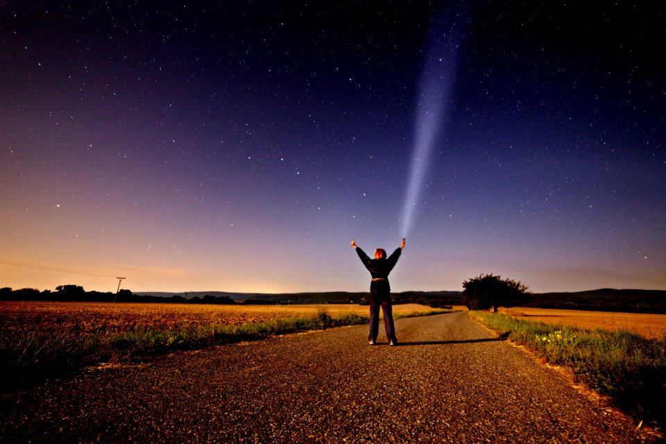 person raising his two hands while holding flashlight standing on road between grass during sunset