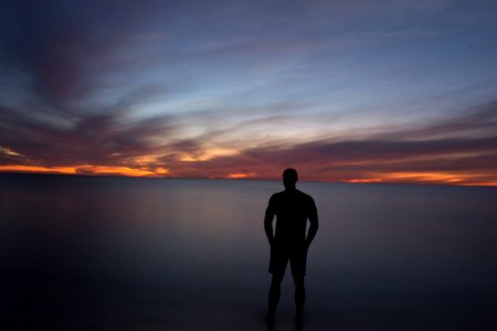 silhouette of person standing near calm body of water during golden hour photo