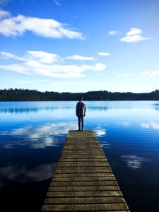 man standing on lake dock watching water under blue sky during daytime photo