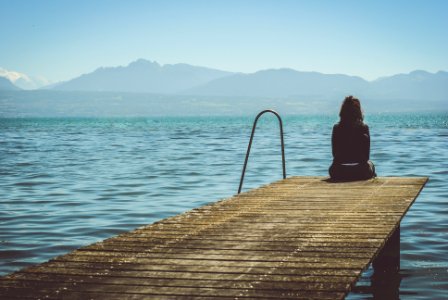 a woman sits on the end of a dock during daytime staring across a lake photo