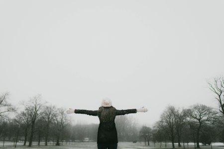 woman in black long-sleeved shirt standing and facing trees with stretched arms under white sky