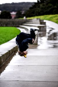 person wears black jacket and black pants doing flip on gray concrete walkway with water during daytime selective focus photography photo
