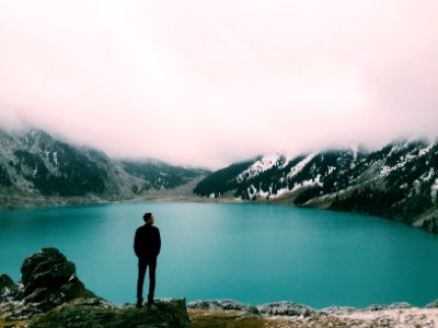 man standing on mountain looking at lake under white clouds at daytime photo