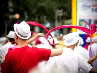 selective focus photography of man wearing pink t-shirt and white fedora hat holding pink ribbon with stick waving on the air during daytime photo