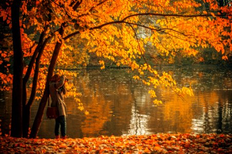 woman in brown coat standing near orange leafed tree and body of water during daytime photo