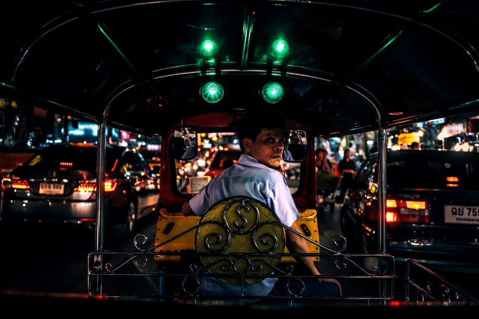 man in white collared shirt sitting in yellow and black autorickshaw between cars with taillights turned on at night
