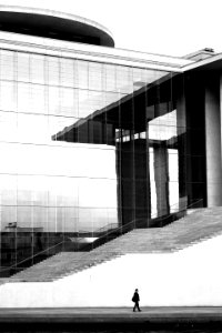 grayscale photo of man walking on concrete pavement beside high building photo