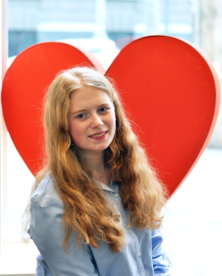 A blonde woman standing in front of a large red heart.