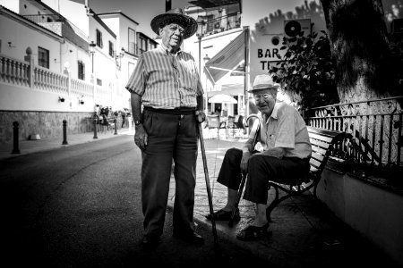 man standing beside a man sitting on cast iron bench near buildings photo