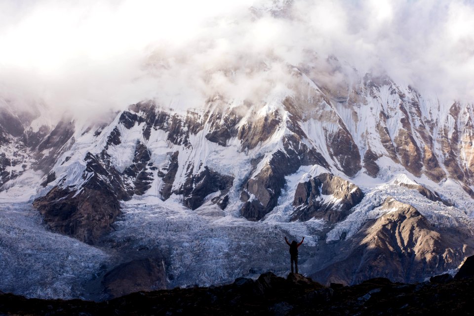 silhouette photo of a person raising hands on top of mountain near gray mountain covered with snow
