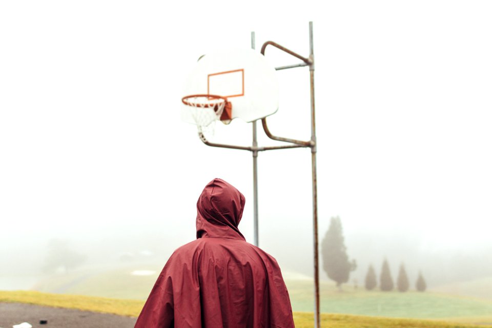 person wearing maroon raincoat standing under white and grey basketball hoop during foggy daytime