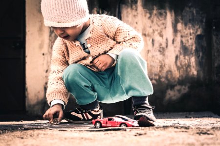 child squatting on beige floor holding rock near red sports coupe scale model at daytime